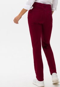 Pantalons stretch bourgogne avec une coupe ajustée, taille mi-haute et deux poches arrière, associés à des baskets blanches et une chemise à manches partiellement retroussées.
