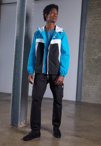 Veste colorée en bleu, noir et blanc, tissu léger, fermeture éclair à l'avant, poignets élastiques ; portée avec un pantalon cargo noir et des baskets noires.