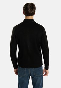 Svart tröja med hög krage, stickad struktur och ribbat avslut. Bärs med blå jeans som visar en slät bakvy.