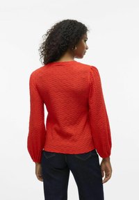 Rote strukturierte Bluse mit puffartigen langen Ärmeln, Rundhals-Ausschnitt und taillierter Passform, kombiniert mit dunklen Jeans. Keine sichtbaren Verzierungen.