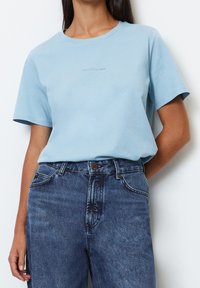 Light blue cotton t-shirt with a round neckline and short sleeves. Features subtle logo print in the center. Paired with dark denim jeans.