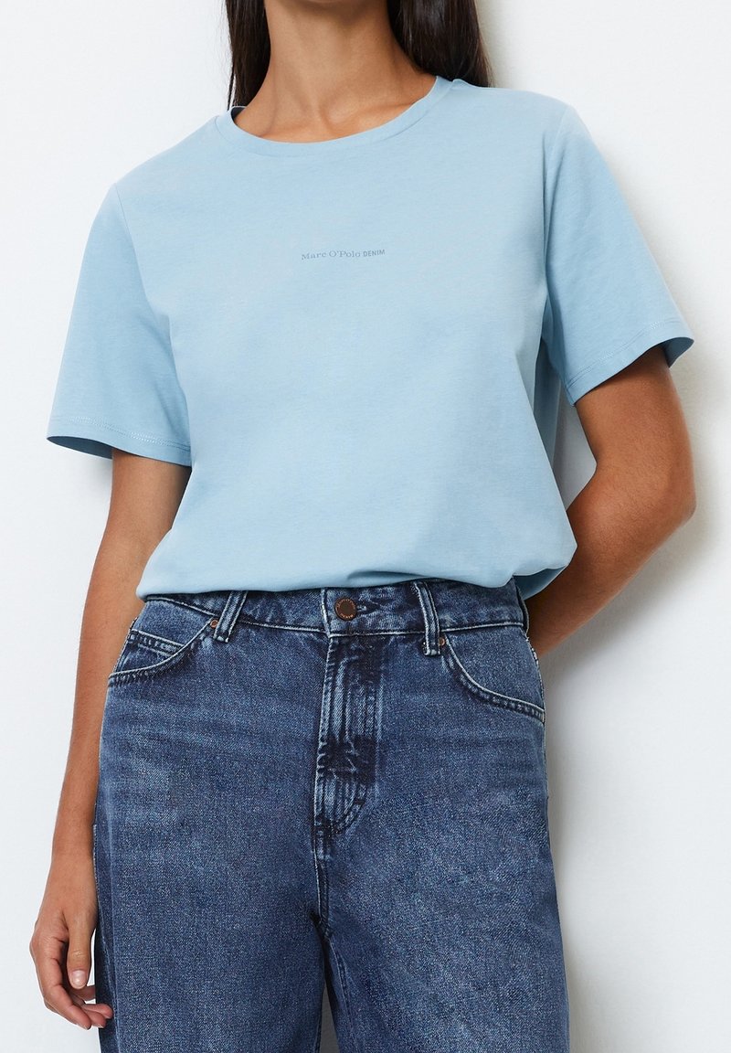 Light blue cotton t-shirt with a round neckline and short sleeves. Features subtle logo print in the center. Paired with dark denim jeans.