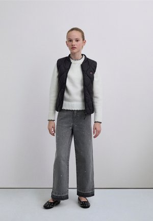 Veste matelassée noire portée sur un pull blanc en maille avec un col côtelé, associée à un jean large noir orné d'accents perle et des chaussures slip-on noires.