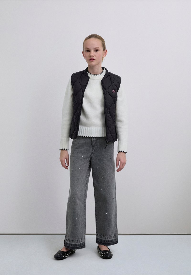 Musta tikattu liivi valkoisen neulotun villapaidan päällä, jossa on ribbineuloskaulus, yhdistetty leveisiin mustiin farkkuihin, joissa on helmikoristelu, ja mustiin slip-on-kengillä.