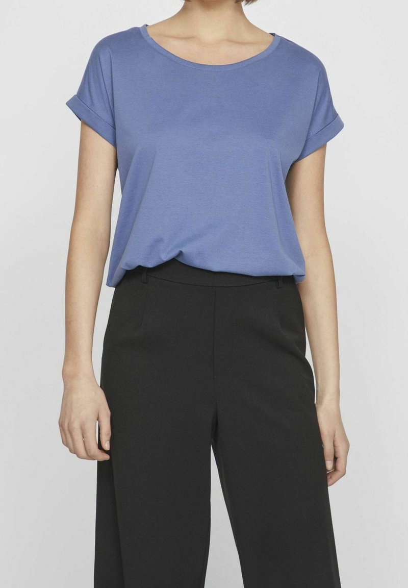 Femme portant un haut bleu à manches courtes rentré dans un pantalon noir taille haute sur un fond uni.