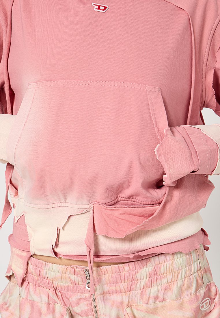 Rosa, kurzes Sweatshirt mit einer Fronttasche, fransigen Kanten und einem Logo. Kombiniert mit rosa Batik-Shorts mit einem elastischen Bund.