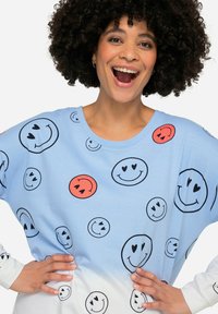 Sudadera de gran tamaño en azul claro con varios patrones de caras sonrientes en negro y naranja, con un degradado a blanco en el dobladillo.