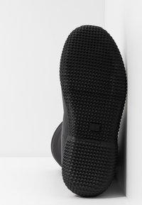 Semelle de chaussure en caoutchouc noir avec un motif texturé en forme de grille, affichée verticalement sur fond blanc.