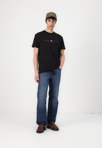 Schwarzes Baumwoll-T-Shirt mit Logo-Druck, kombiniert mit ausgeblichenen blauen Jeans. Braune Schuhe mit leichtem Glanz und einer karierter Mütze.
