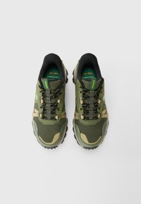 Zöld és bézs hálós sneaker pár fekete és sárga cipőfűzőkkel, fekete bélésekkel és masszív talppal, előre nézve egy fehér felületen.
