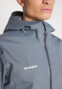 Grau wasserfeste Jacke mit hohem Kragen, Frontreißverschluss und hellgrauem Innenfutter. Die Markenbezeichnung "MAMMUT" ist in Weiß auf der Brust aufgedruckt.