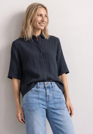 Femme souriante aux cheveux blonds porte une chemise noire texturée à boutons et un jean bleu clair, debout devant un fond clair uni.