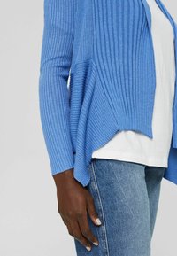 Mörkhyad person som bär en blå ribbad cardigan över en vit skjorta och blå jeans, med handen vilande vid sidan.