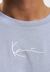 Lyseblå t-shirt med ribbet halsudskæring og et hvidt broderet logo på forsiden. Blød tekstur med en glat finish.
