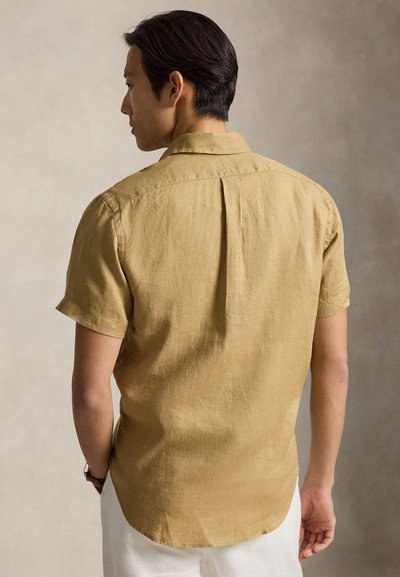 Άνδρας με κοντομάνικο λινό πουκάμισο μουσταρδί και λευκό παντελόνι, φαίνεται από πίσω σε ουδέτερο φόντο.