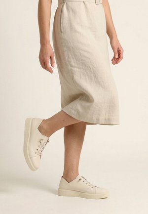 Partie inférieure du corps portant une jupe midi beige et des baskets crème à lacets, un pied légèrement levé du sol.