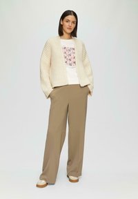 Cremfarbener gerippter Cardigan über einem weißen T-Shirt mit geometrischem Muster, kombiniert mit locker sitzenden beigen Hosen und beigen Turnschuhen.