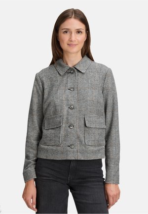 Vrouw in een grijs geruit knopenjack met twee voorzakken en zwarte broek, staand tegen een effen lichte achtergrond.