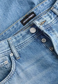 Lyseblå denimjeans med en sort mærkat med "JACK&JONES", sølvknapper og subtilt slidtekstur på stoffet.