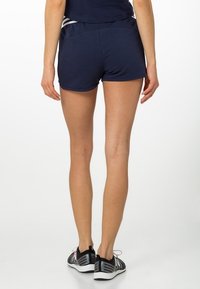 Marina atletiska shorts med en slät yta, elastisk midja och reflekterande silvriga detaljer. Kombineras med svarta sportskor.