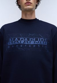 Sweat-shirt bleu marine avec un imprimé graphique texturé mettant en avant "NAPAPIJRI" et "geographic" dans des tons de bleu plus clair. Design classique avec col rond.