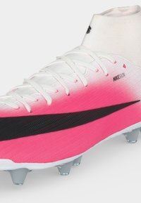 Λευκό και ροζ ποδοσφαιρικό παπούτσι Nike με μαύρο σήμα Swoosh, σχεδιασμό ψηλής κάλτσας στον αστράγαλο, λευκά κορδόνια και μεταλλικά studs στη σόλα.