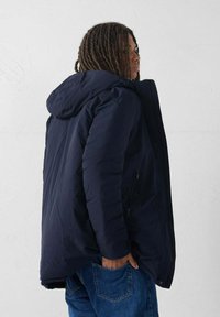 Persona con dreadlocks di lunghezza media che indossa una giacca a cappuccio blu navy e jeans blu, in piedi di lato con una mano in tasca.