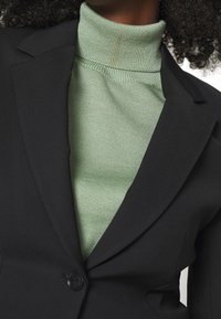 Donna che indossa un blazer nero sopra un maglione a collo alto a coste verde chiaro, con capelli ricci parzialmente visibili.