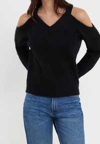 Pull en maille noire avec design épaules dénudées, col en V et manches longues. La texture semble douce, associé à un jean en denim bleu.