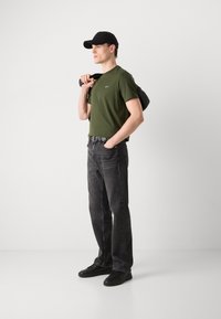 Zelené bavlněné tričko s malým logem, černé džíny, černé tenisky a černá kšiltovka. Model nese černou tašku přes jedno rameno.