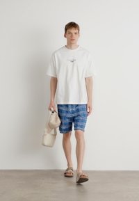 Camisa de algodón blanca con un logo, pantalones cortos de cuadro azul y bolso beige. El modelo lleva chanclas marrones. Fondo minimalista.