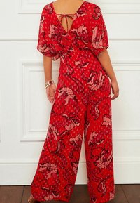 Jumpsuit floral rouge avec des jambes larges et fluides. Doté d'une fermeture à nouer à l'arrière et de manches courtes en tête de chauve-souris. Tissu texturé avec des accents roses.