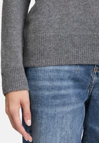 Linke Hand einer Person in hellgrauem Strickpullover und blauer Jeans, die an ihrer Seite gegen einen neutralen Hintergrund ruht.