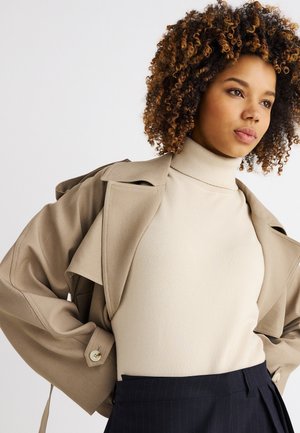 Kvinde med krøllet hår iført en beige trenchcoat over en cremefarvet rullekrave og mørke bukser med tynde striber, der ser eftertænksomt til siden.