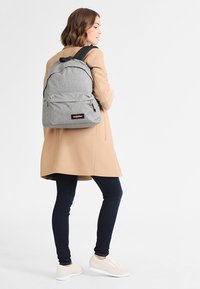 Grauer Stoffrucksack mit verstellbaren schwarzen Gurten, vorderer Reißverschlusstasche und Markenlogo. Getragen über einem beigen Mantel und dunklen Jeans.