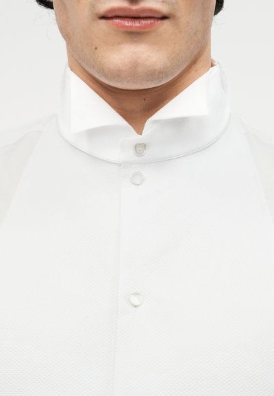 Άνδρας που φοράει λευκό πουκάμισο με γιακά, κουμπωμένο μέχρι πάνω, με ορατό το κάτω μέρος του προσώπου και το λαιμό, σε απλό φόντο.