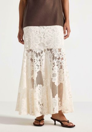 Donna che indossa una gonna midi in pizzo color crema con motivi floreali e sandali con tacco marrone scuro, in piedi davanti a uno sfondo neutro.