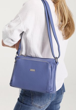 Nainen, jolla on valkoinen teksturoitu paita ja siniset farkut, kantaa teksturoitua sinistä nahkaista olkalaukkua, jossa on etutaskun vetoketju.