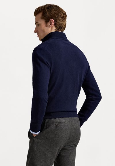 Navy blue sweater with a high collar, knit texture, and ribbed cuffs. Paired with gray trousers featuring a back pocket.