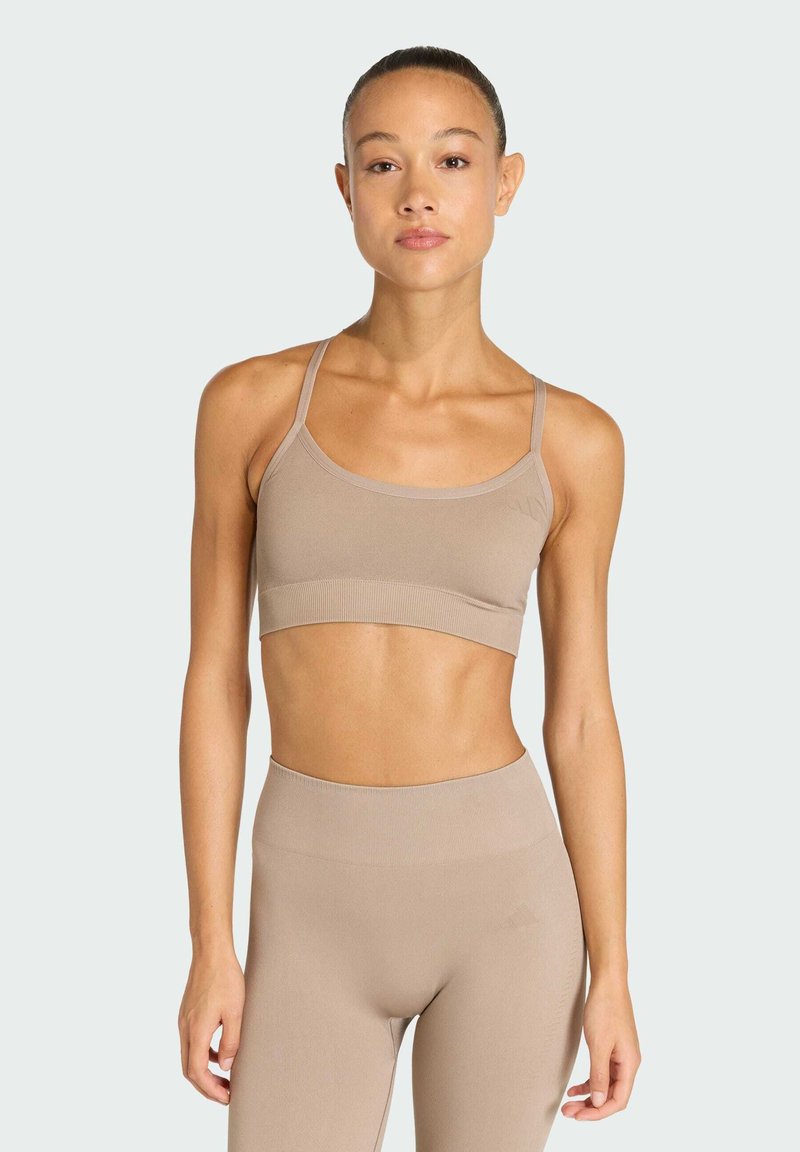 Žena nosící béžový bezšvový sportovní podprsenku a legíny s vysokým pasem, stojící s uvolněnými pažemi proti jednobarevnému světlému pozadí.