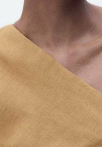 Gelbes Leinengewebe mit einem diagonalen Schnitt, das eine glatte Textur und ein leicht zerknittertes Erscheinungsbild am Hals- und Schulterbereich aufweist.