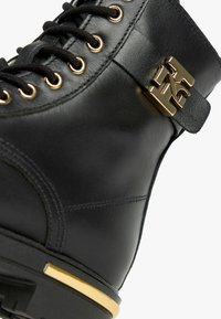 Botte en cuir noir avec ferrures dorées, présentant des lacets ronds, une bride latérale et un talon accentué d'or. Texture lisse et design structuré.