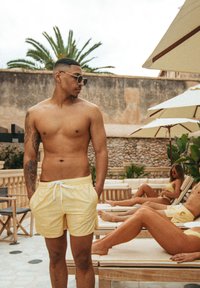 Pantaloni da mare gialli con cordoncino bianco, indossati da un uomo con un tatuaggio, in piedi vicino a lettini prendisole. Due donne in costumi da bagno gialli coordinati sullo sfondo.