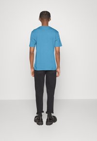 Mężczyzna stojący plecami, ubrany w niebieską koszulę z krótkim rękawem, czarne zrolowane spodnie i czarne masywne buty, na jednolitym tle.