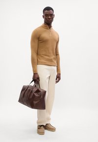 Borsa in pelle marrone con doppio manico, pantaloni color crema e una maglietta a maniche lunghe color tan con zip a metà. Calzature casual.