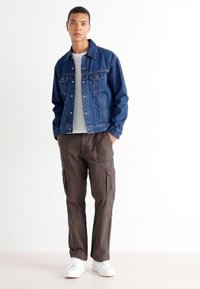 Donkerdenimjack met knopen aan de voorkant, wit t-shirt en bruin cargobroek met meerdere zakken, aangevuld met witte sneakers.