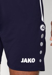Shorts sportivi navy con branding bianco "JAKO" e motivi circolari accentuati sui lati. Realizzati in tessuto liscio e elastico.