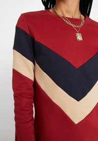 Sweatshirt rouge et marine avec un motif en chevron beige, col rond, manches longues et texture douce. Inclus un collier en chaîne dorée et un pendentif.