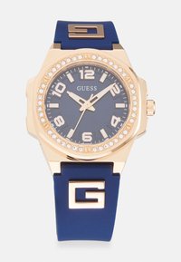 Reloj dorado con correa de silicona azul. Presenta una esfera azul oscura, acentos dorados y un bisel adornado con cristales. Monograma en la correa.