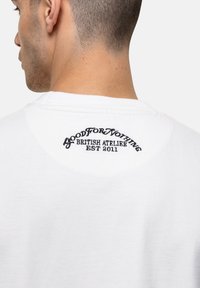 Witte katoenen t-shirt met een ronde halslijn en zwarte geborduurde tekst op de achterkant met de tekst "GOOD FOR NOTHING BRITISH ATELIER EST 2011."